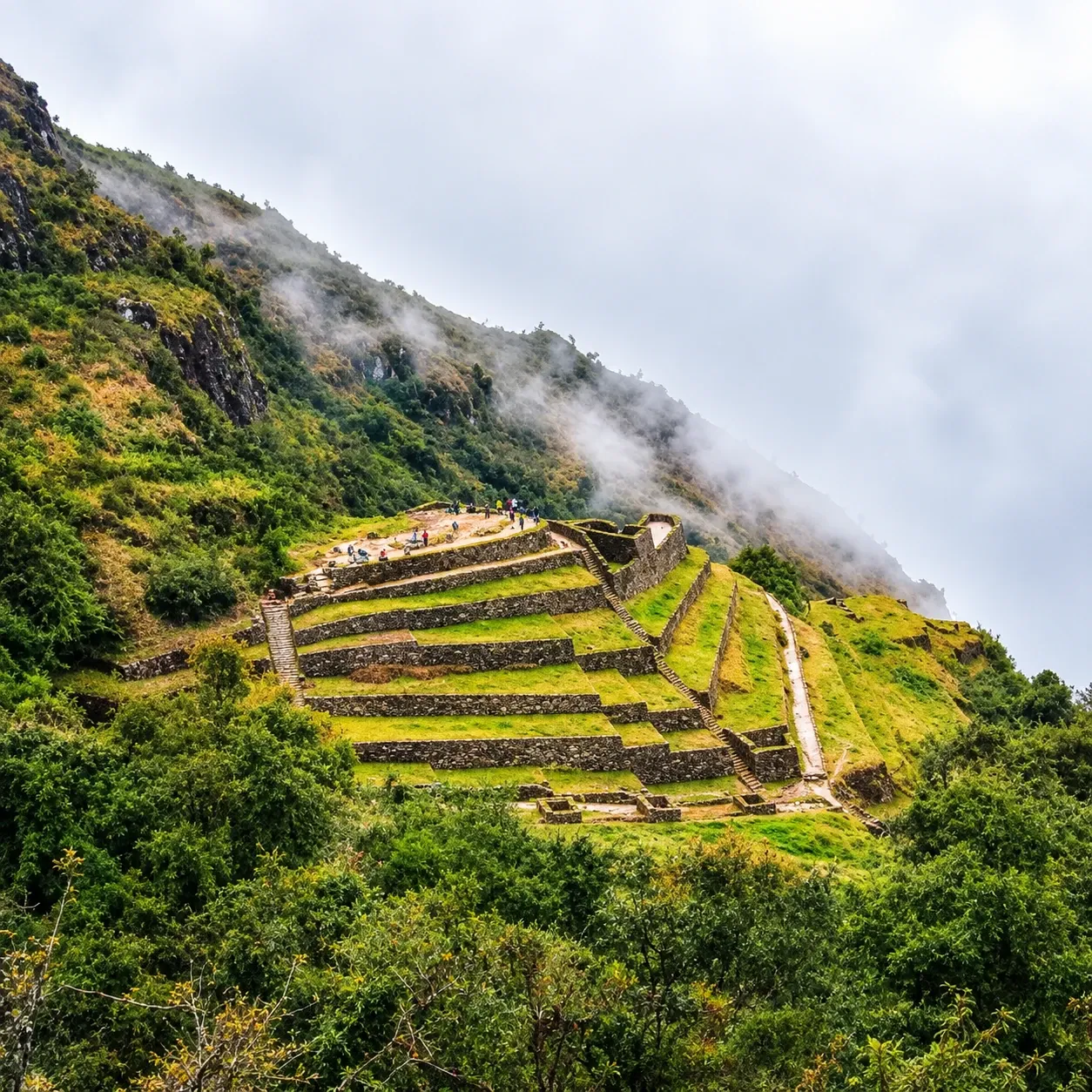 Inca terraces on a steep forested slope, cloud pouring over the ridge