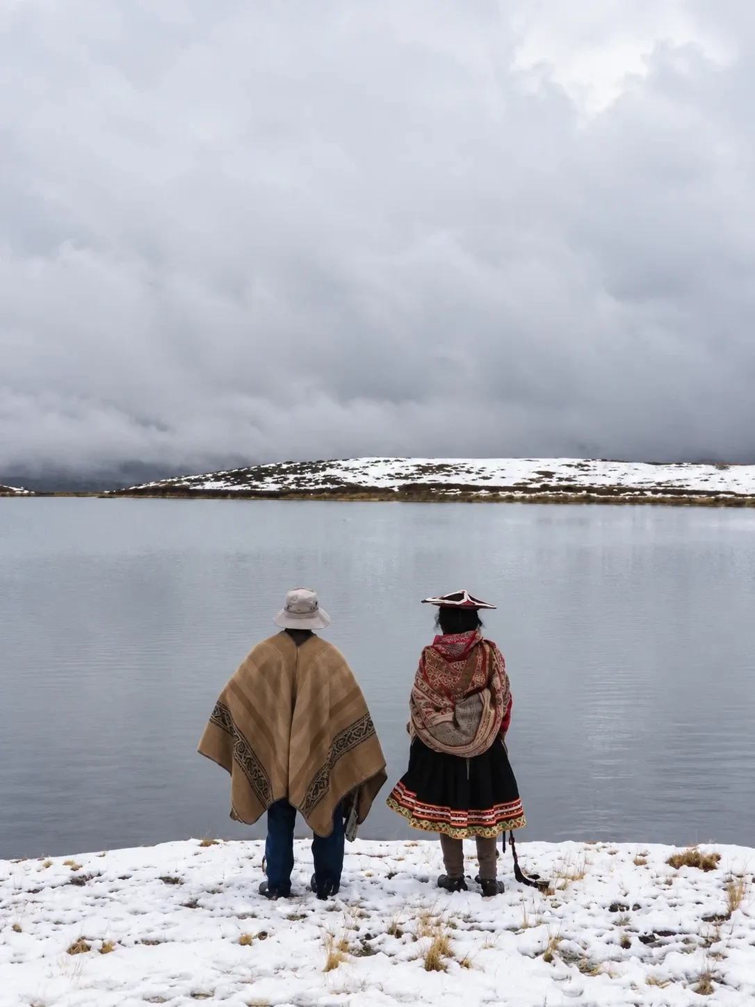 Two people in traditional dress beside a high lake after snowfall